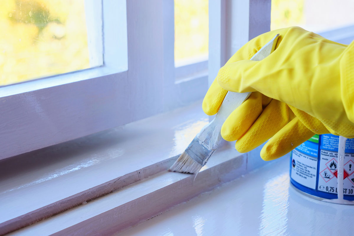 Hand in yellow rubber gloves applies a glossy paint finish to window casement