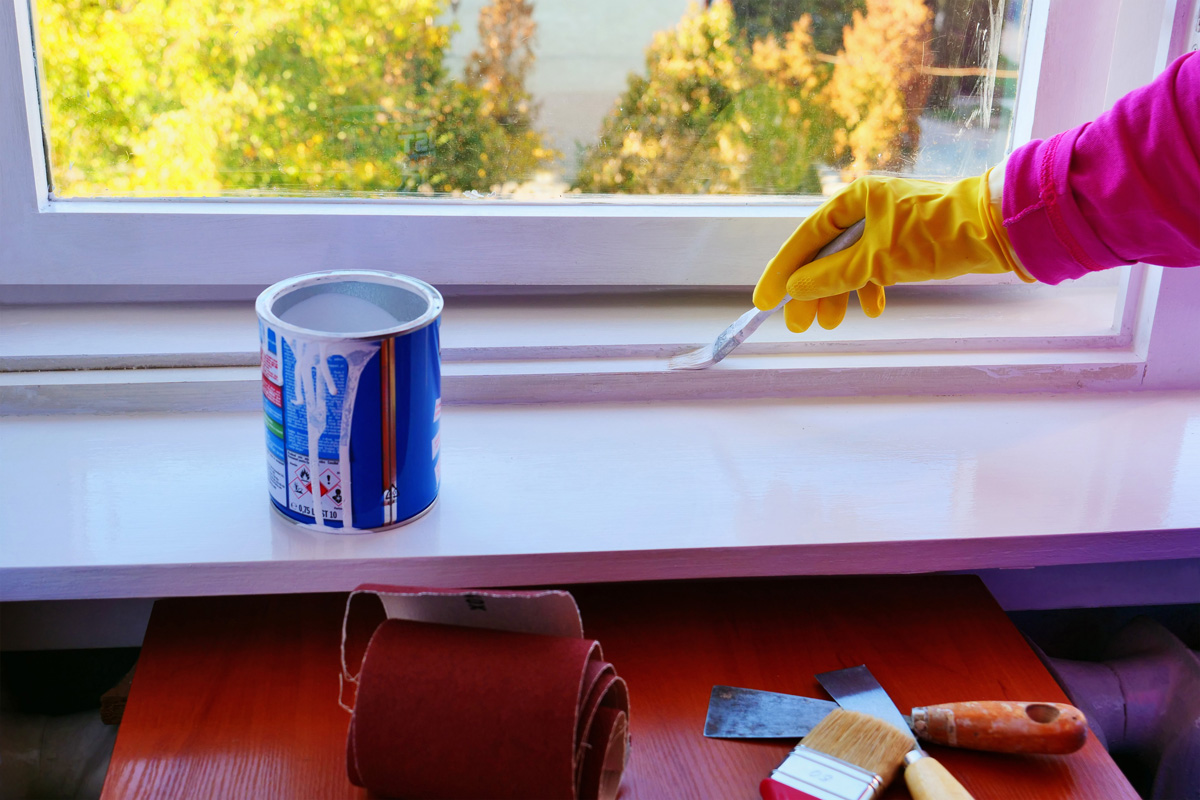 Hand in yellow rubber gloves applies a glossy white paint finish to a window frame.