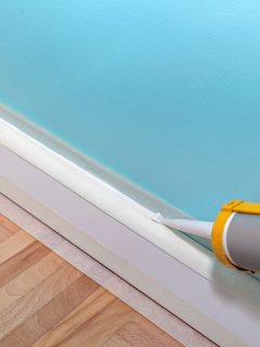 Mans hand caulk skirting board with caulking gun and silicone cartrige Filling gaps in base board using masking tape, How To Caulk Baseboards To Floor [Steps Explained]