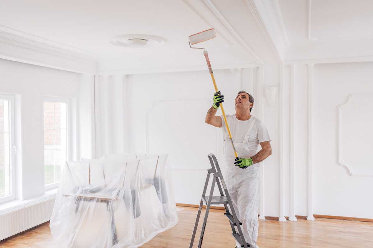 One man, mature male house painter standing on a ladder and painting a ceiling with paint roller, indoors in a house.