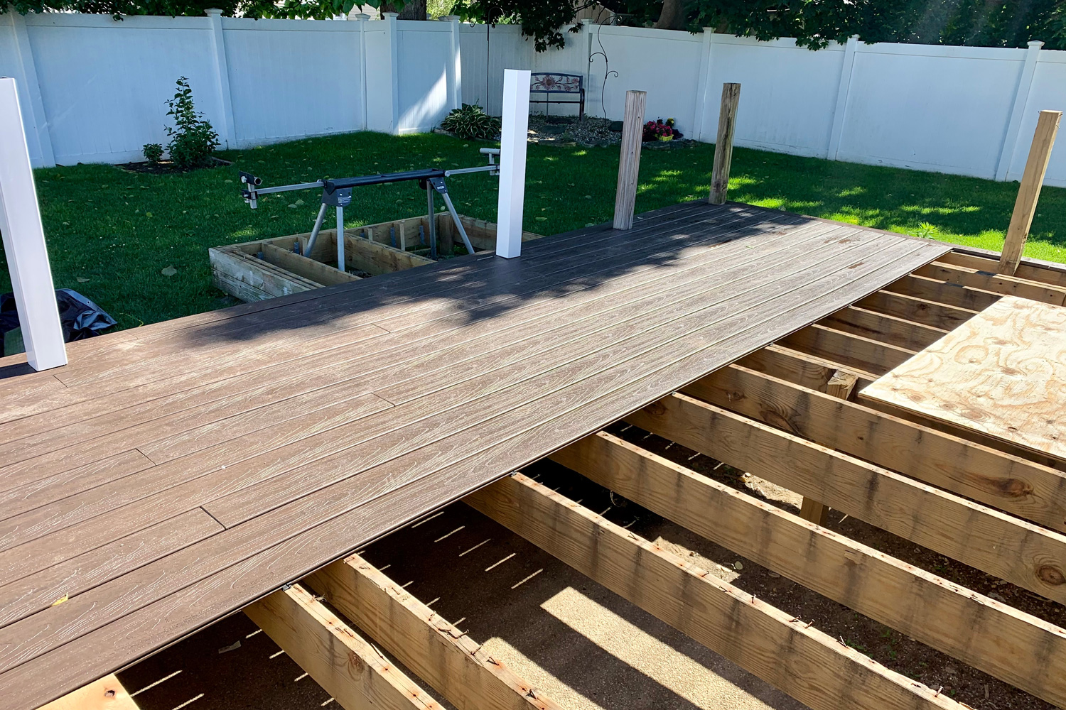 Remodeling a deck in a suburban backyard. The contractor is laying down composite wood , has once PVC railing in place, and there is a giant saw. The tool is sitting in the demolished deck.