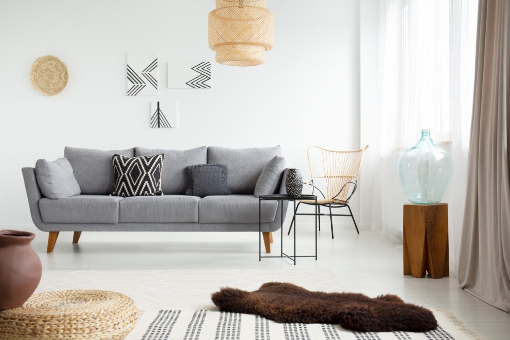 Real photo of a brown, fur rug lying on white floor in front of a gray couch with cushions in bright living room interior