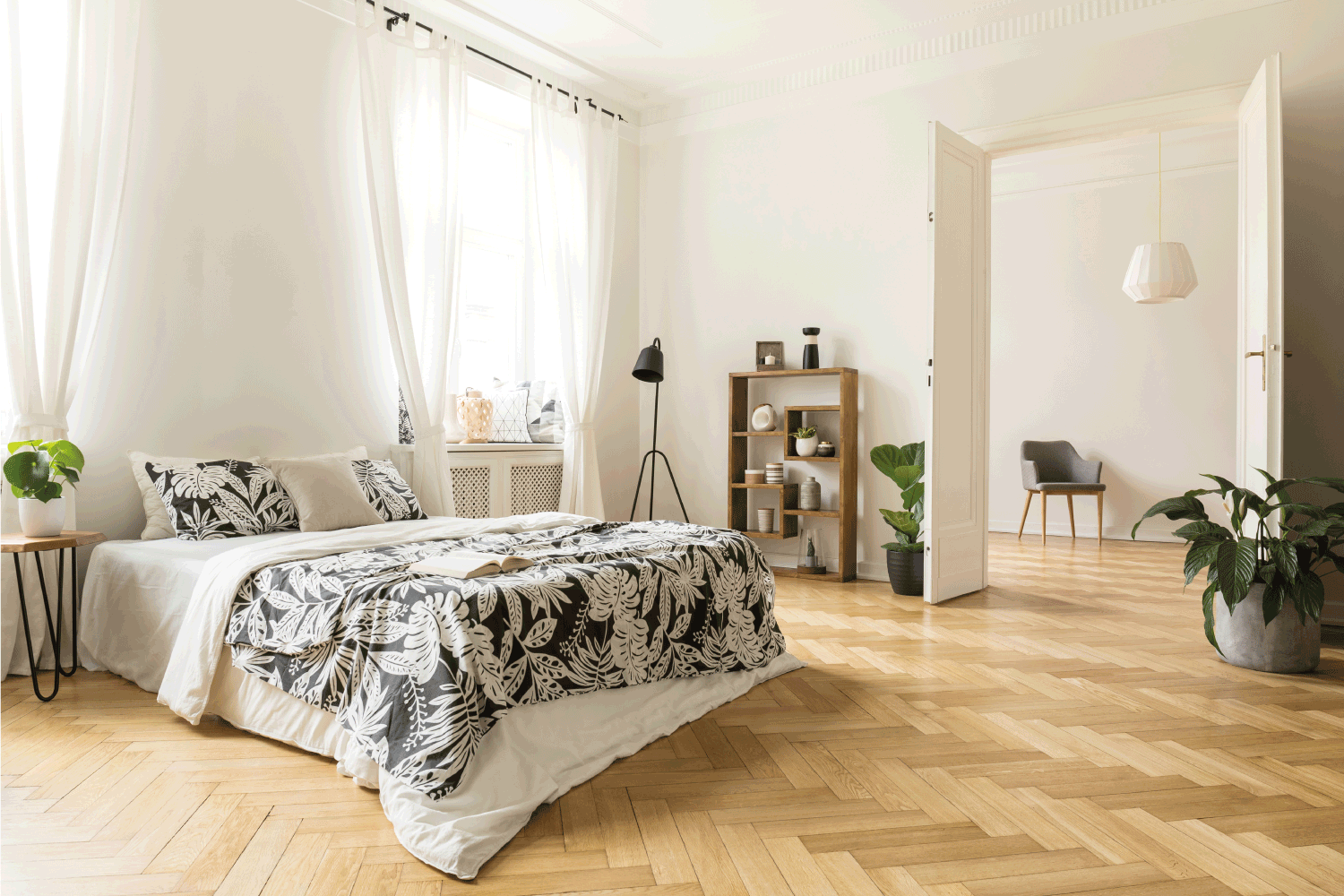 Stylish apartment interior with white walls and herringbone wooden floor. A view from a bedroom with a big bed to another room with an armchair