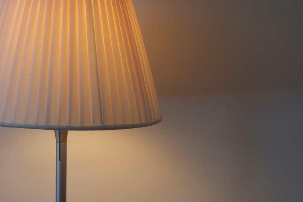 Table lamp with pleated linen shade. Paint texture of wall visible.