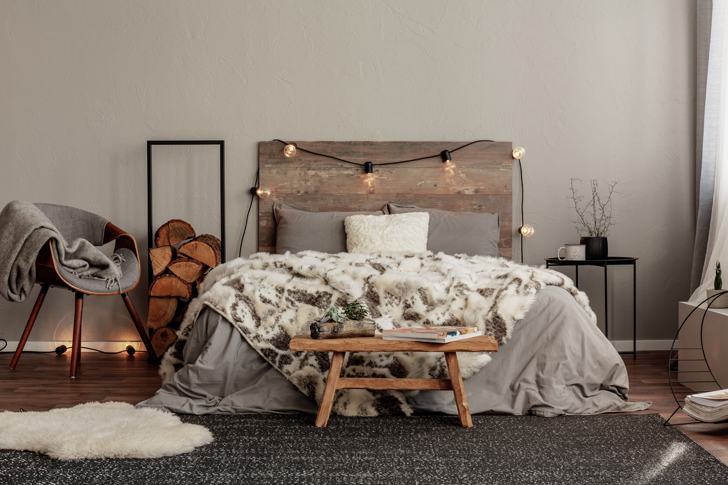 Trendy grey chair with blanket and log of wood next to cozy double bed with wooden headboard and light bulbs