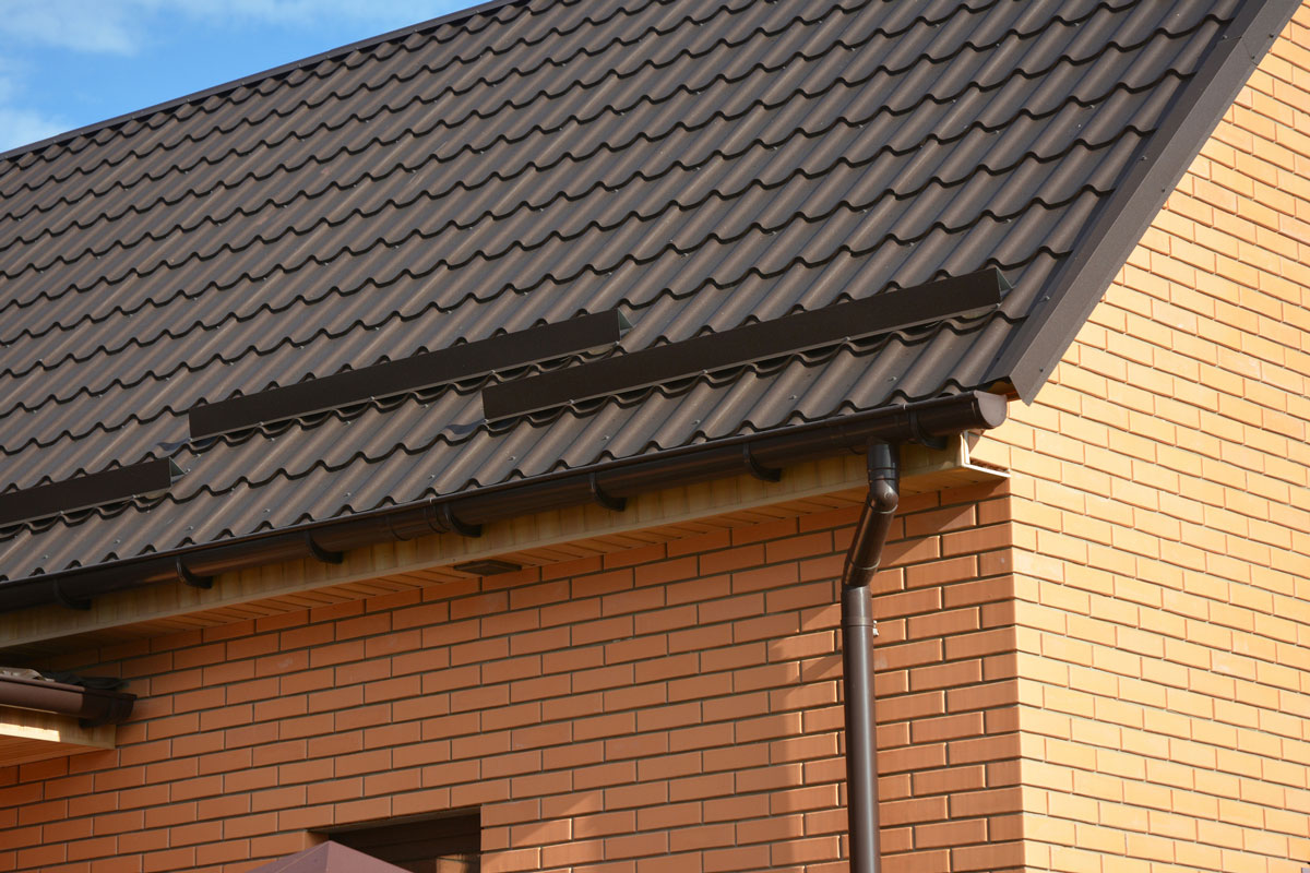 a metal brown roof of a brick house with snow stoppers, snow guards, a rain gutter, a downspout, soffit and fascia board