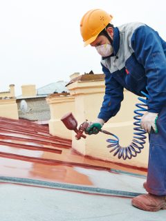 roofer builder worker with pulverizer spraying paint on metal sheet roof - How Long Does Metal Roof Paint Last