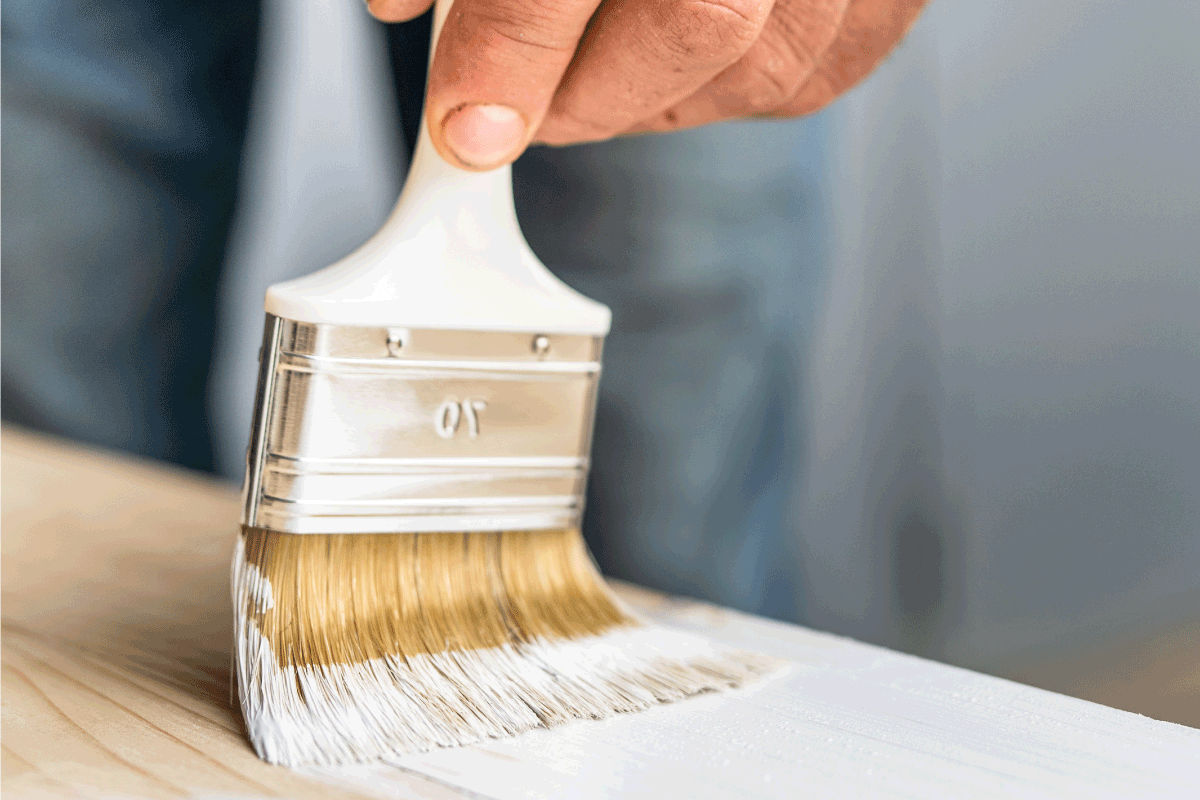 white paint brush in the hand of unknown man painting wooden board. How To Whitewash Laminate Furniture