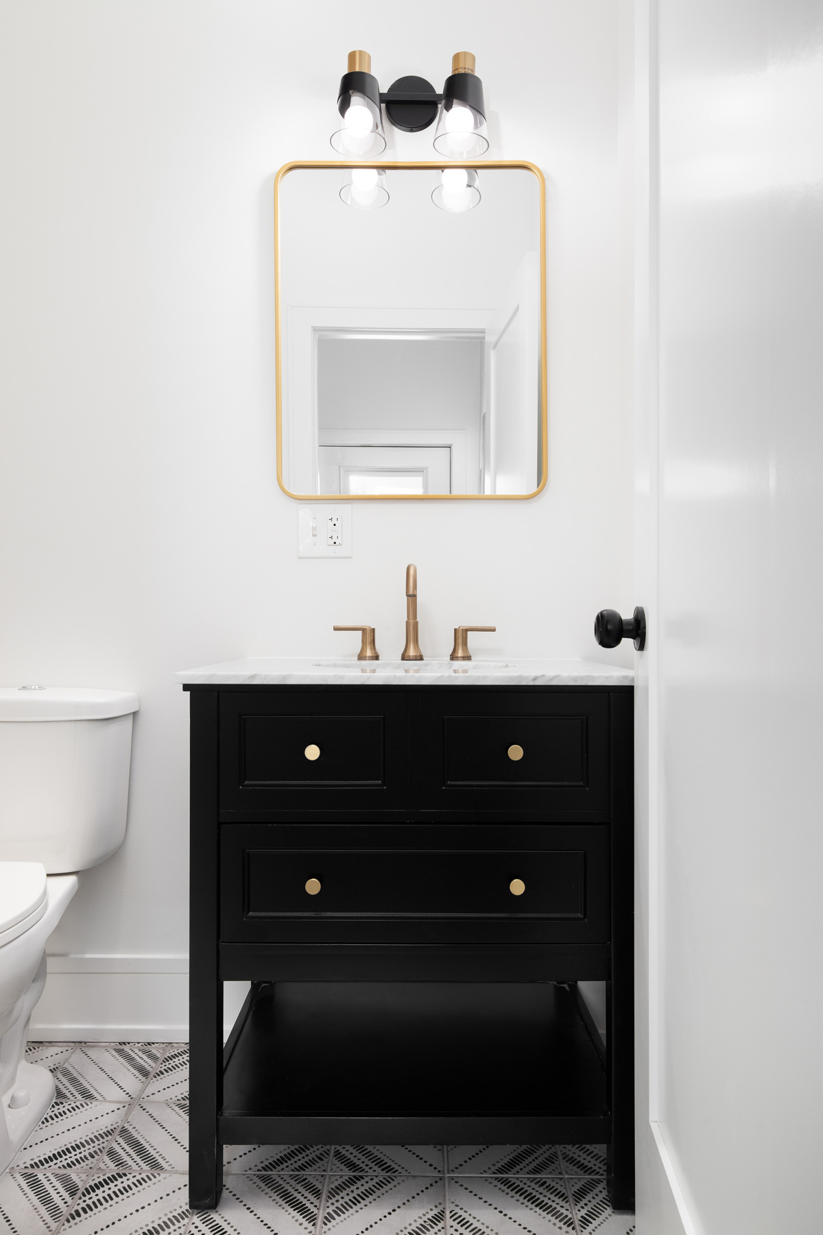 A bathroom with a black vanity, gold faucet and mirror, and a mosaic tiled floor.