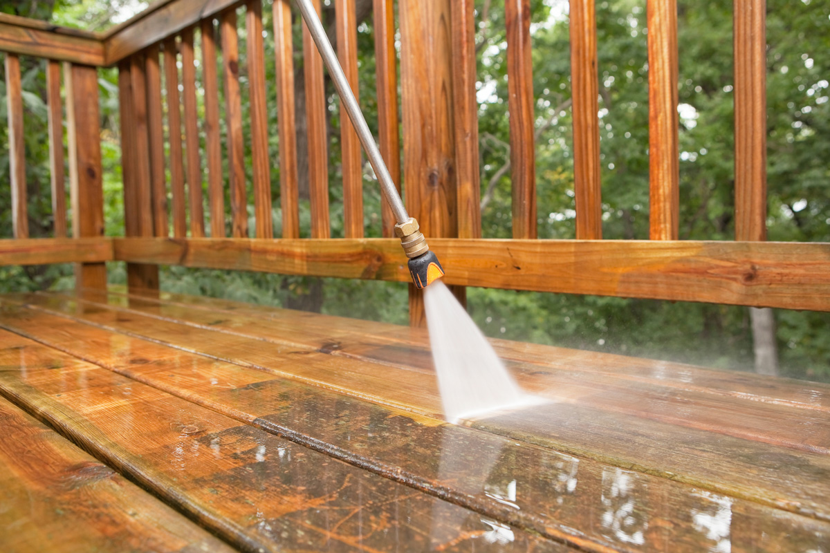 A pressure washer sprayer is cleaning a weathered treated wood deck.