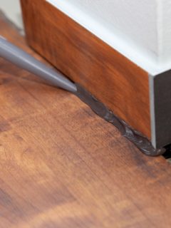 Applying caulk on the baseboard inside wooden floored living room, Should You Caulk Between Floor And Baseboard?