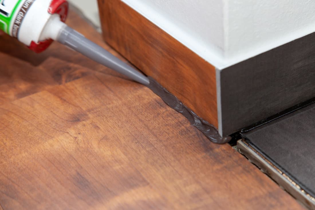 Applying caulk on the baseboard inside wooden floored living room