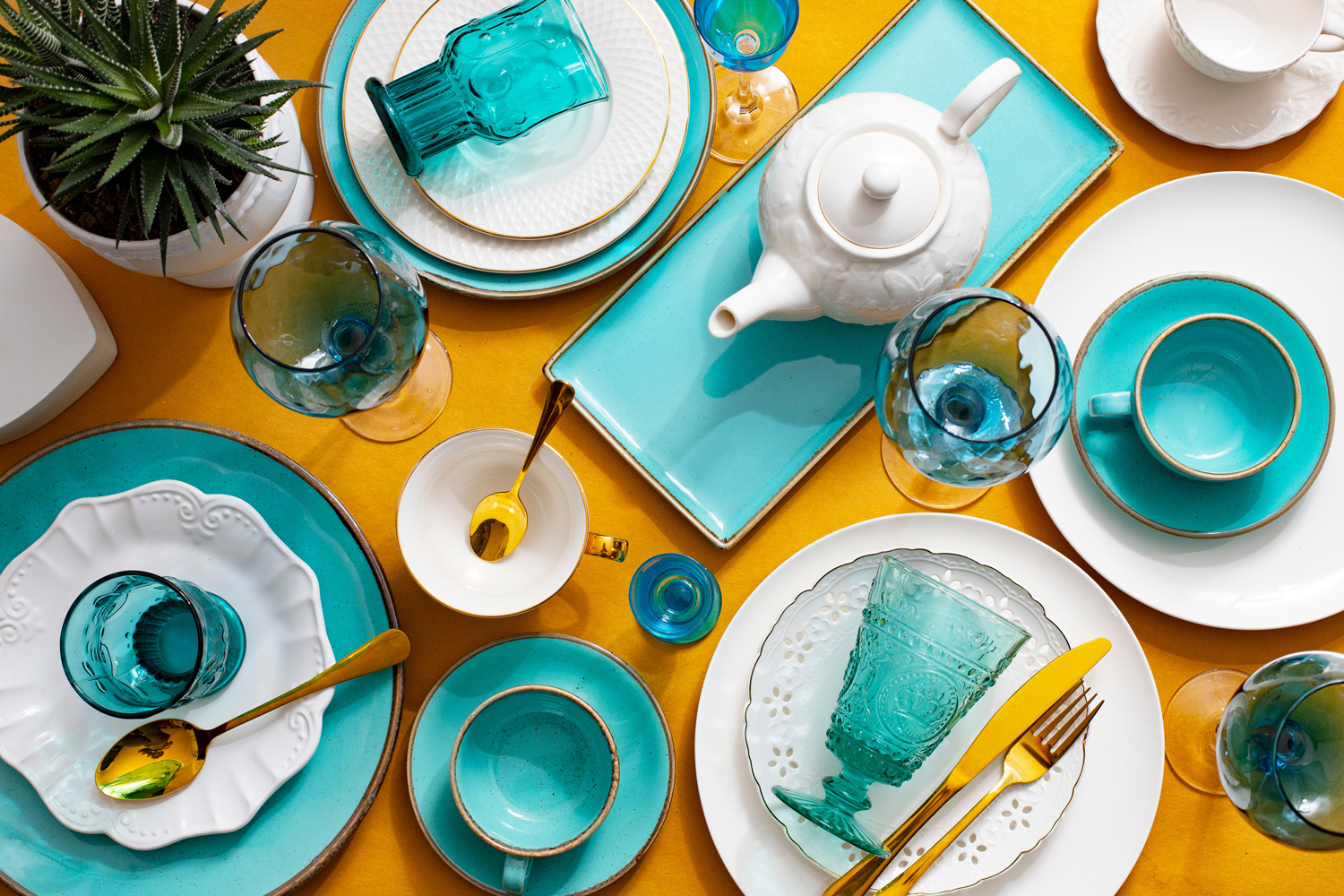 Beautiful table setting with colourful blue green dishes, cups, glasses and other dishnware on a mustard yellow background