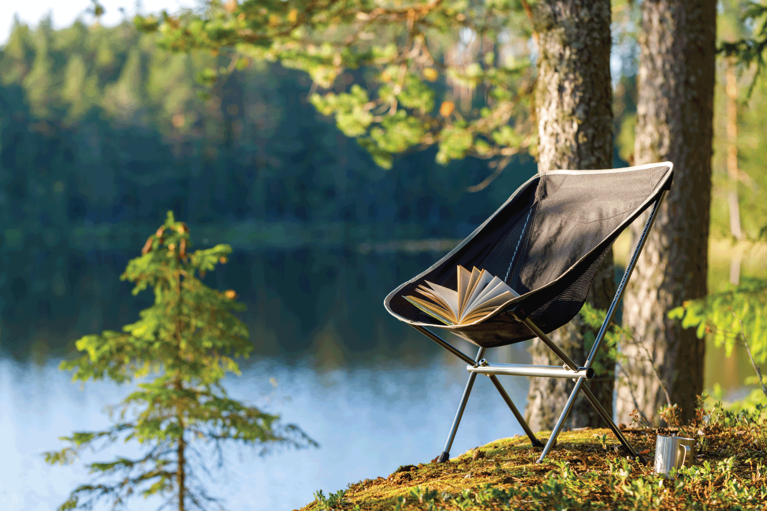 Camping folding chair with a book and a mug on the background of a forest lake.