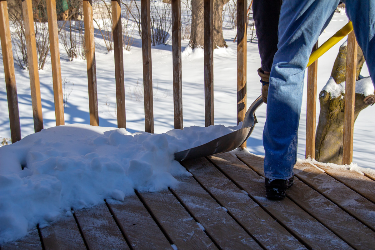 This image shows a partial view of an unidentified adult person shoveling deep snow off of a wooden deck on a sunny day.