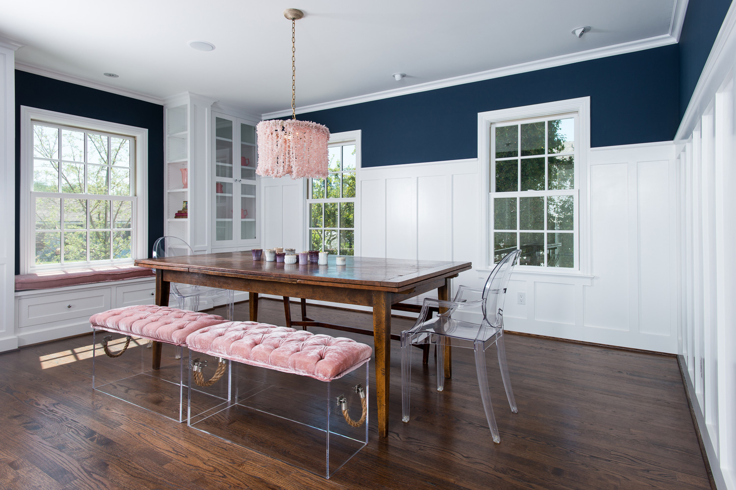 Dining room with distressed country wood table and lucite chairs and bench. The room has hard wood flooring, painted wood wainscoting and cabinets. The upper wall has a dark navy flat paint and the ceiling has adjustable recessed lights.