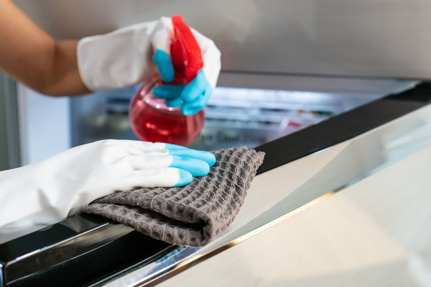 Housewife hands in rubber protective glove with microfiber cloth and detergent spray cleaning a refrigerator at home. Closeup