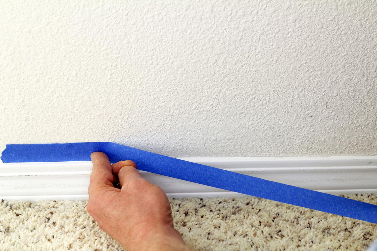 Male hand preparing to paint wall trim by placing blue painter's tape on the wall above it for protection.