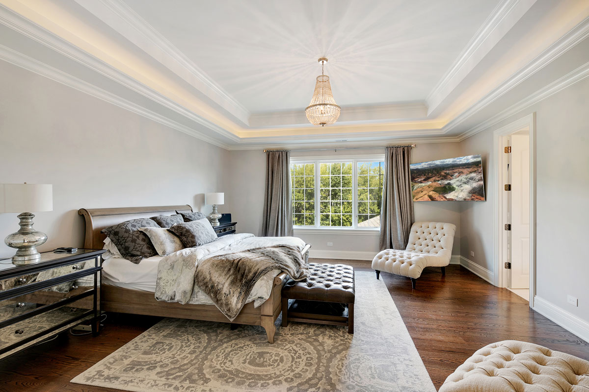 Modern spacious bedroom with tray ceiling, dark hardwood flooring and a gorgeous chandelier hanged on the center