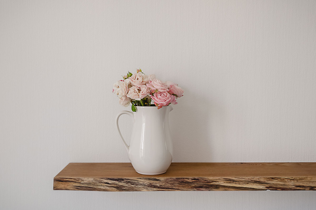 Open floating wooden shelf with a big ceramic jug of pink pastel flowers on a white wallpaper wall in a modern design living room