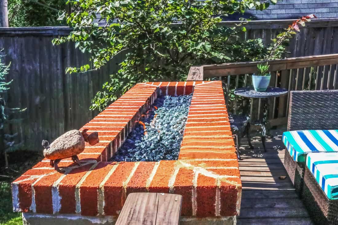 Outdoor natural gas fire pit with low flames on patio with privacy fence in background and colorful seating
