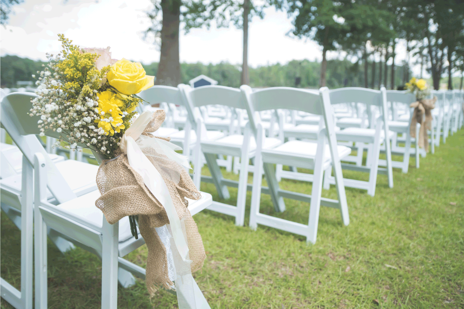 Pink and yellow flower arrangement with burlap and lace ribbon on white folding chair at wedding ceremony