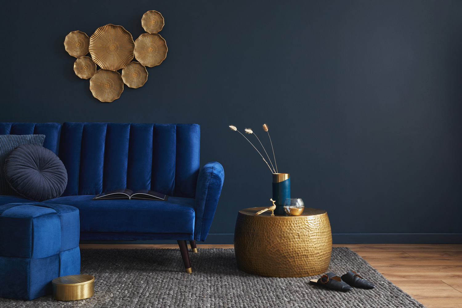Stylish living room interior composition with velvet blue sofa, golden side table, pouf, pillows and elegant home decor. Dark blue wallpaper. Template.