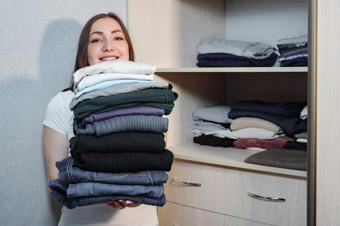 Smiling young woman standing with heap of folded clothing in hands smiling at camera