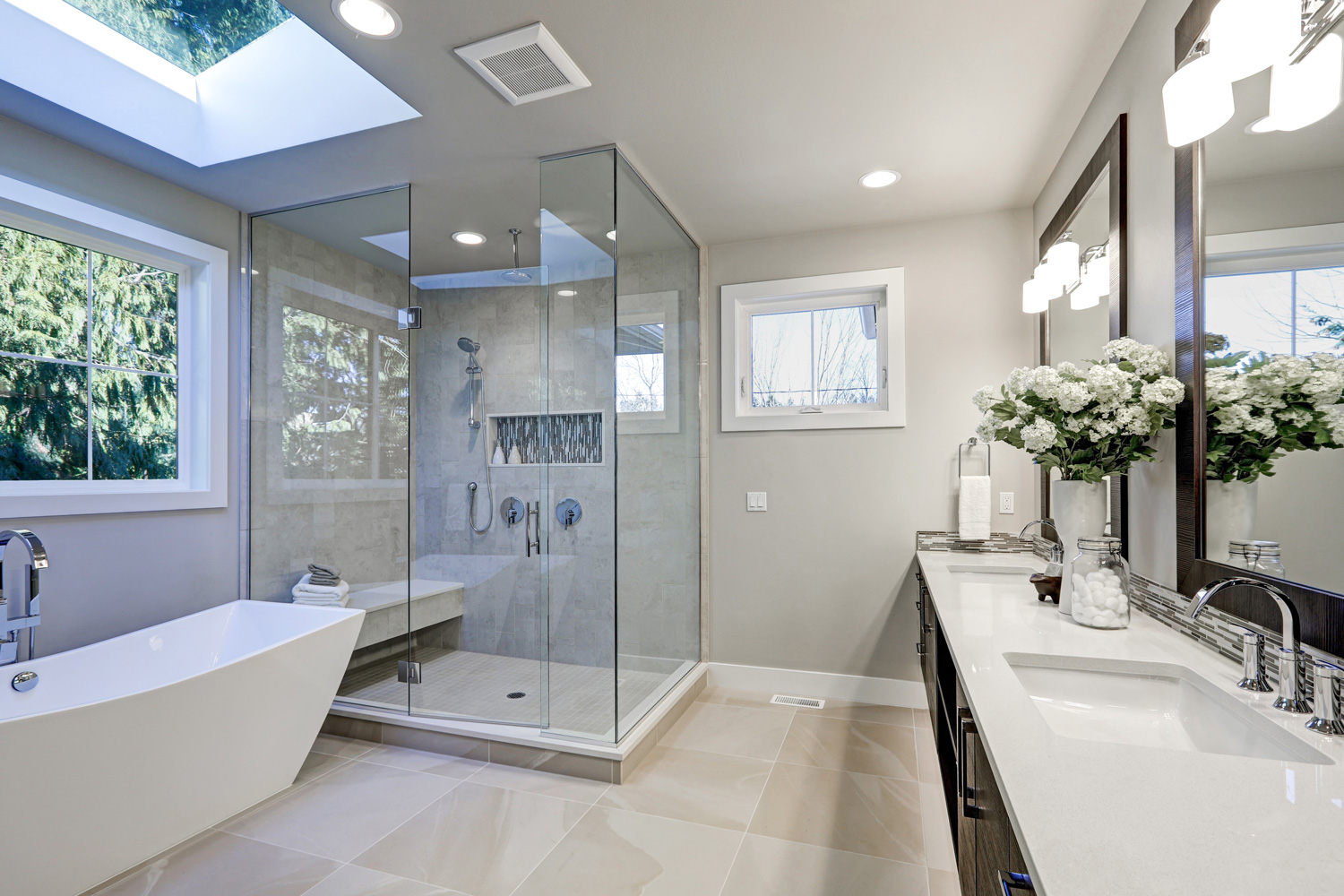 Spacious bathroom in gray tones with heated floors, freestanding tub, walk-in shower, double sink vanity and skylights. 