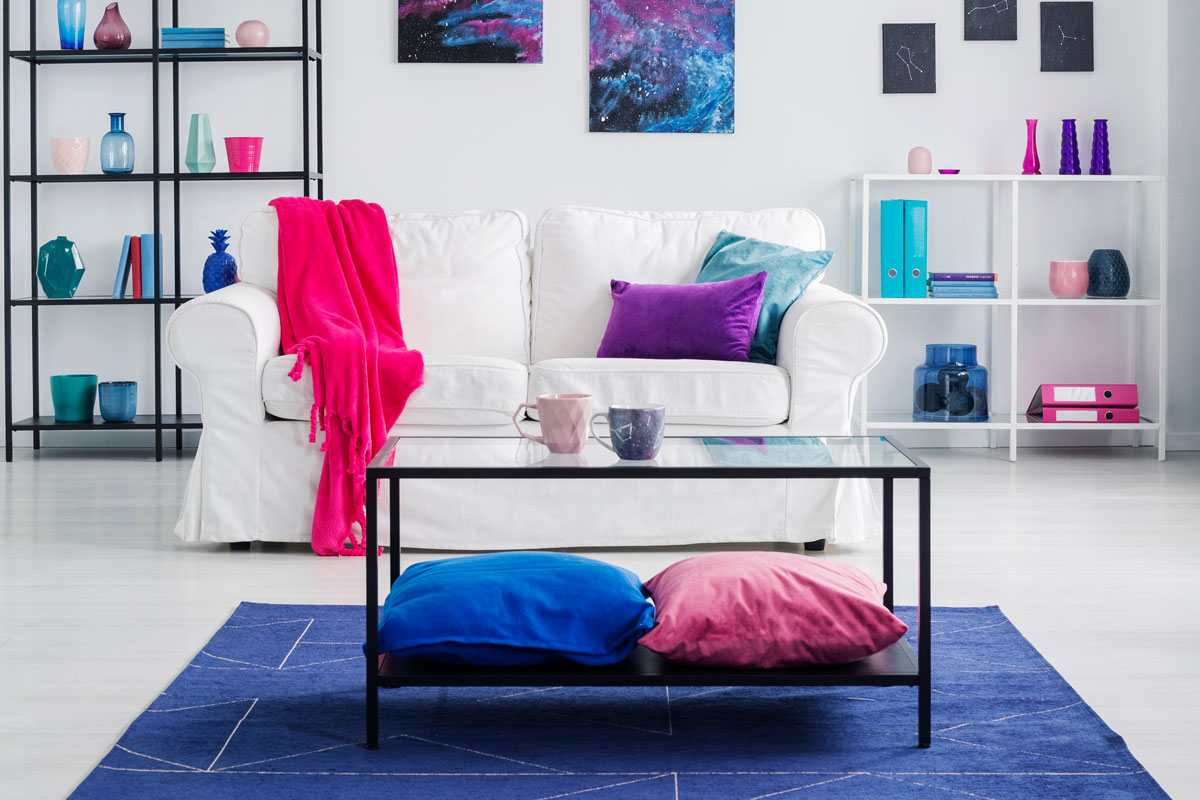 Table with cushions on blue carpet in living room interior with posters above white sofa