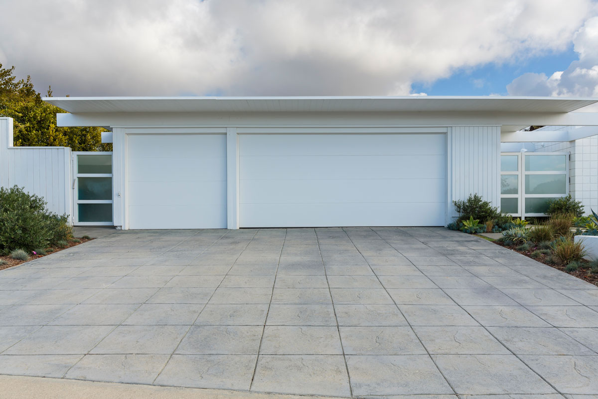 front view of white three car garage door with a long driveway, partly cloudy sky. the house is a mid-century modern style house