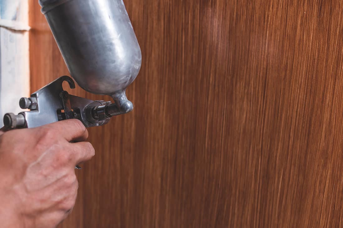 A handyman sprays lacquer wood finish to the end panel of the bottom cabinet in the kitchen