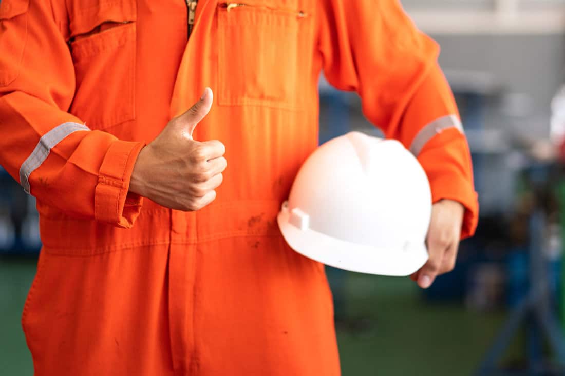 A worker in orange coverall is thumb up action and holding white safety helmet with factory workshop as blurred background.