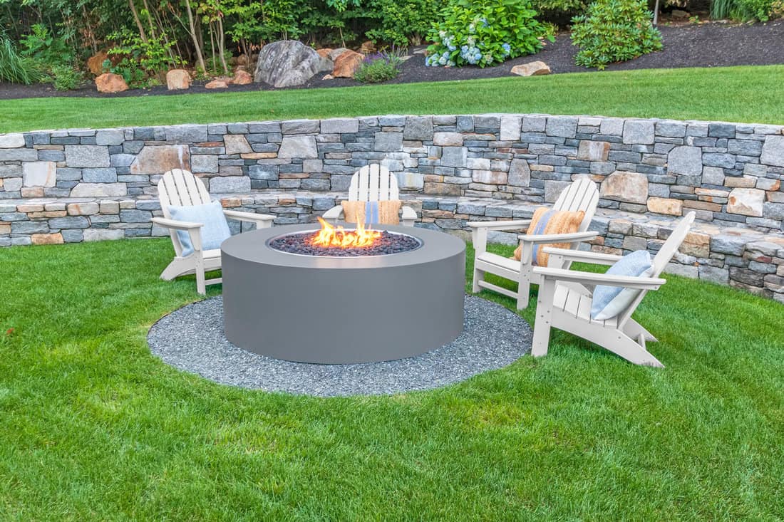 Adirondack chairs around fire pit with stone wall and lush green grass.