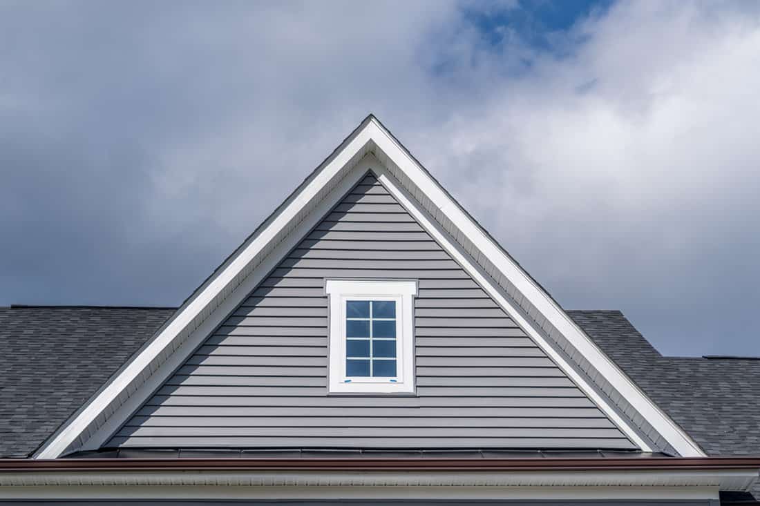 Gable with grey horizontal vinyl lap siding, double hung window with white frame, vinyl shutters on a pitched roof attic at a luxury American single family home neighborhood