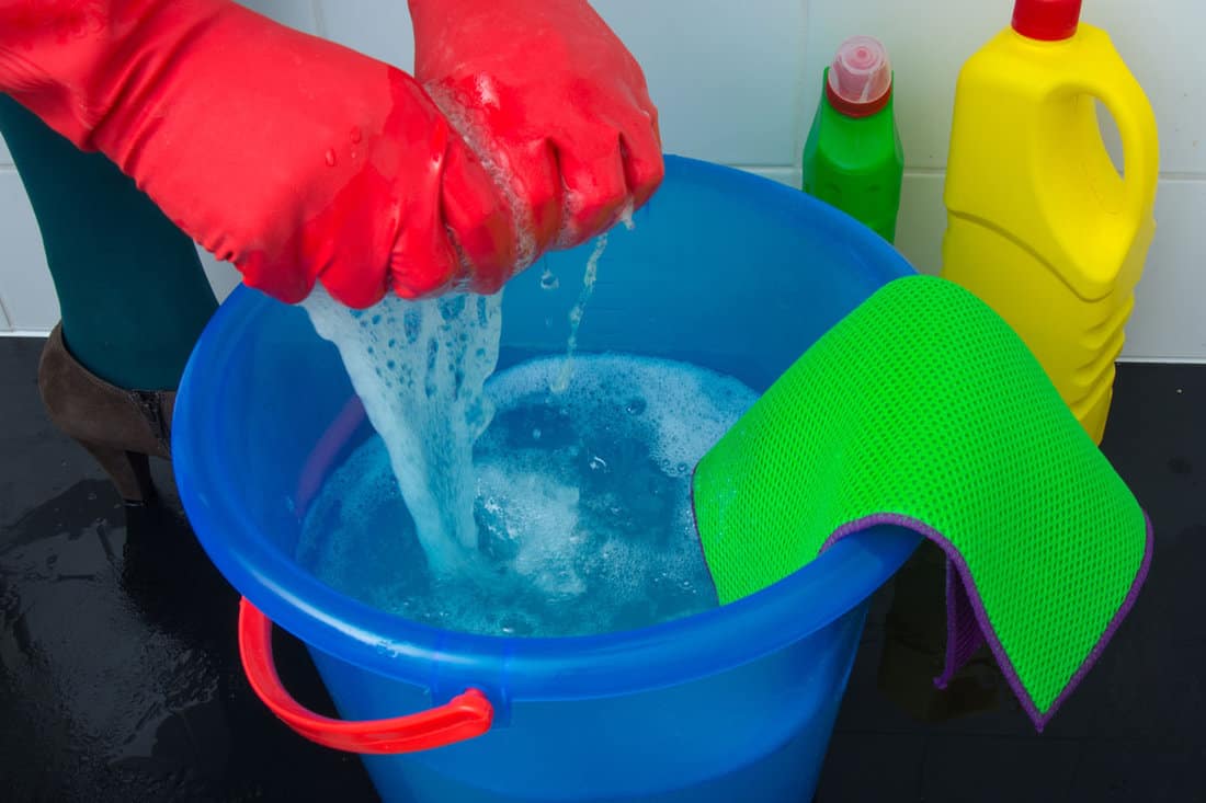Ground level cleaning floor with water and cleaner in a blue bucket woman in red cleaning gloves, green cloth, yellow bleach, sponge