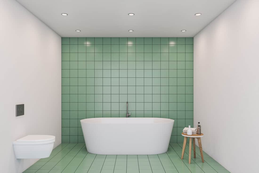 Interior of comfortable bathroom with green tile and white walls, big white bathtub and white toilet. 