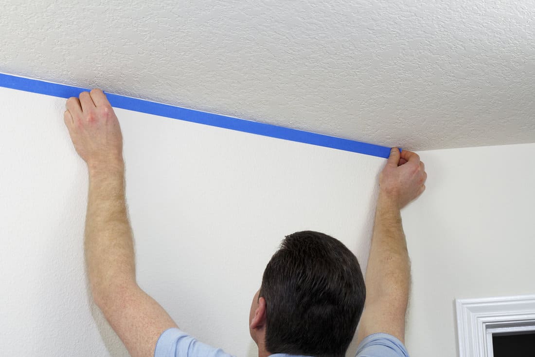 Man preparing to paint ceiling by masking off the wall beneath it with blue painter's tape