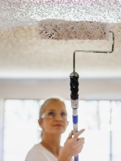 Mature Adult Female Painting Ceiling White With Paint Roller - How To Blend Ceiling Paint To Match Existing Shade