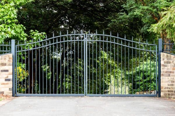 Metal-driveway-property-entrance-gates-set-in-brick-fence-with-garden-trees-in-background