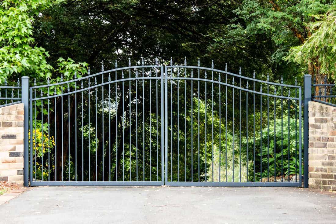 Metal driveway property entrance gates set in brick fence with garden trees in background