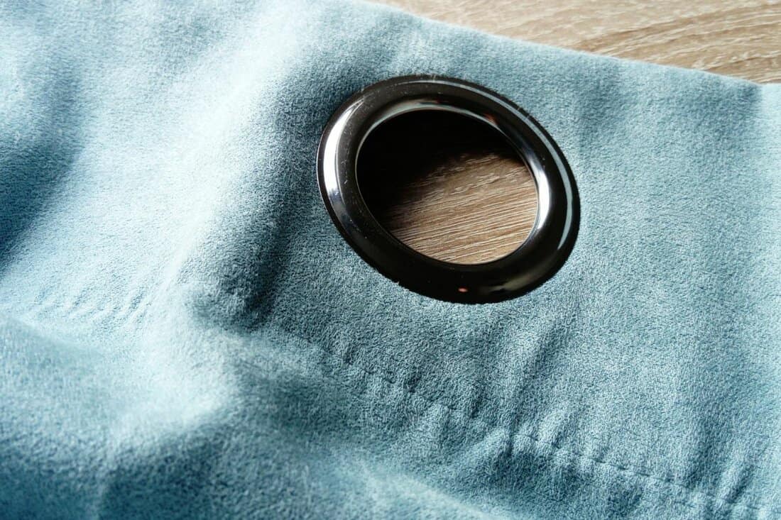 Metal grommet on a gray-blue velvet curtain. The fabric lies on a wooden table. The hole on the material is made of silver coated aluminum. Close-up of a metal eyelet