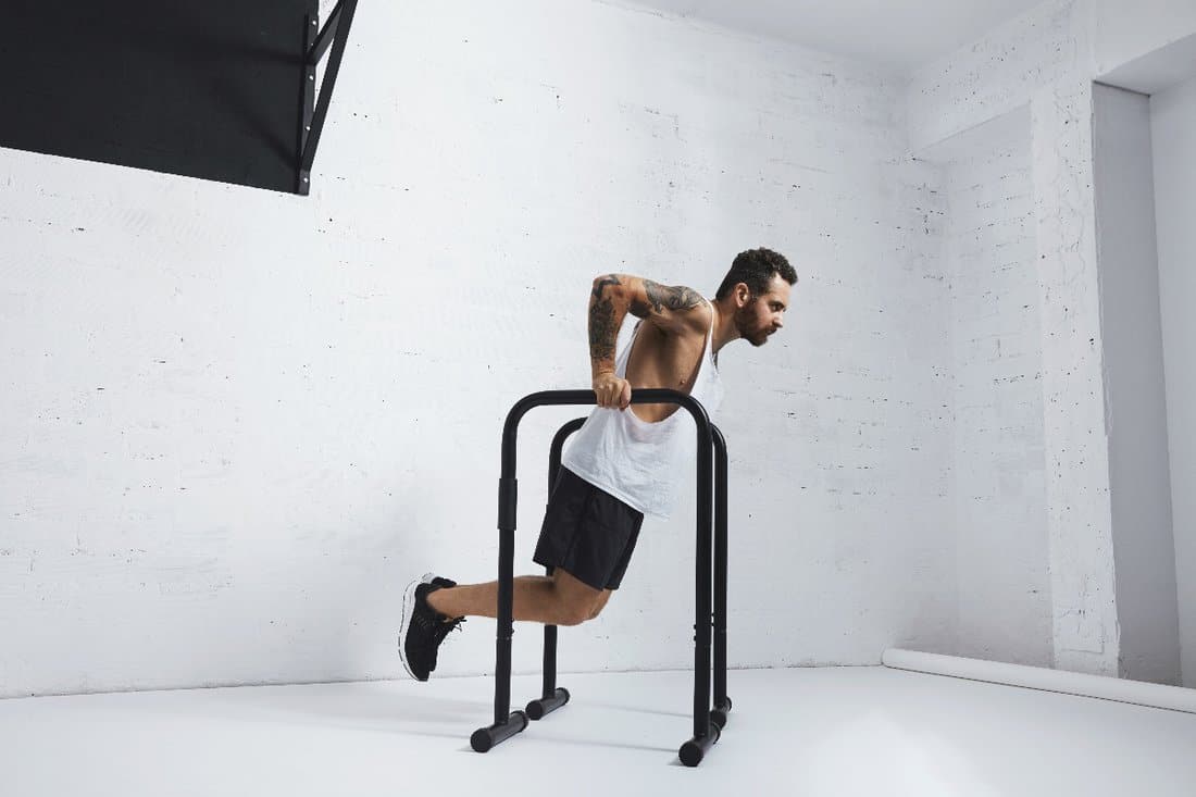 Should You Do Dips Every Day, Strong tattooed in white unlabeled tank t-shirt male athlete shows calisthenic moves Holding dip position on parallel bars