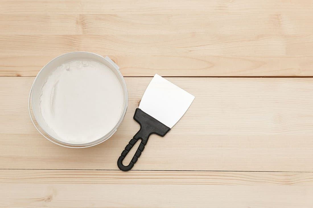 Spatula and a bucket of white putty on wooden boards.