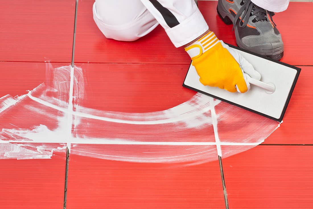 Worker with rubber trowel applying white grout on red tiles on floor