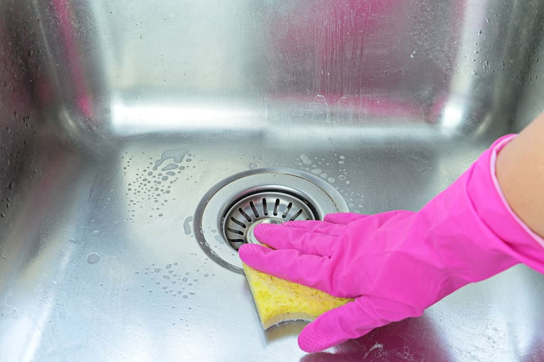 human hand wearing pink washing gloves holding a yellow sponge, cleaning the stainless sink