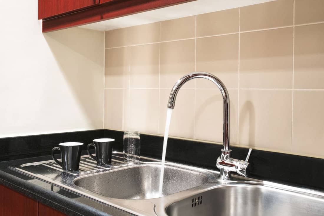 neat modern kitchen stainless sink on the kitchen, beige wall tile, faucet on, water running