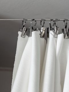 A cream curtain hangs on a rope on a metal hook, against the background of the ceiling and wall, illuminated by daylight - How To Hang Curtains Without A Rod [DIY Creative Ideas For Both Sheers And Curtains]