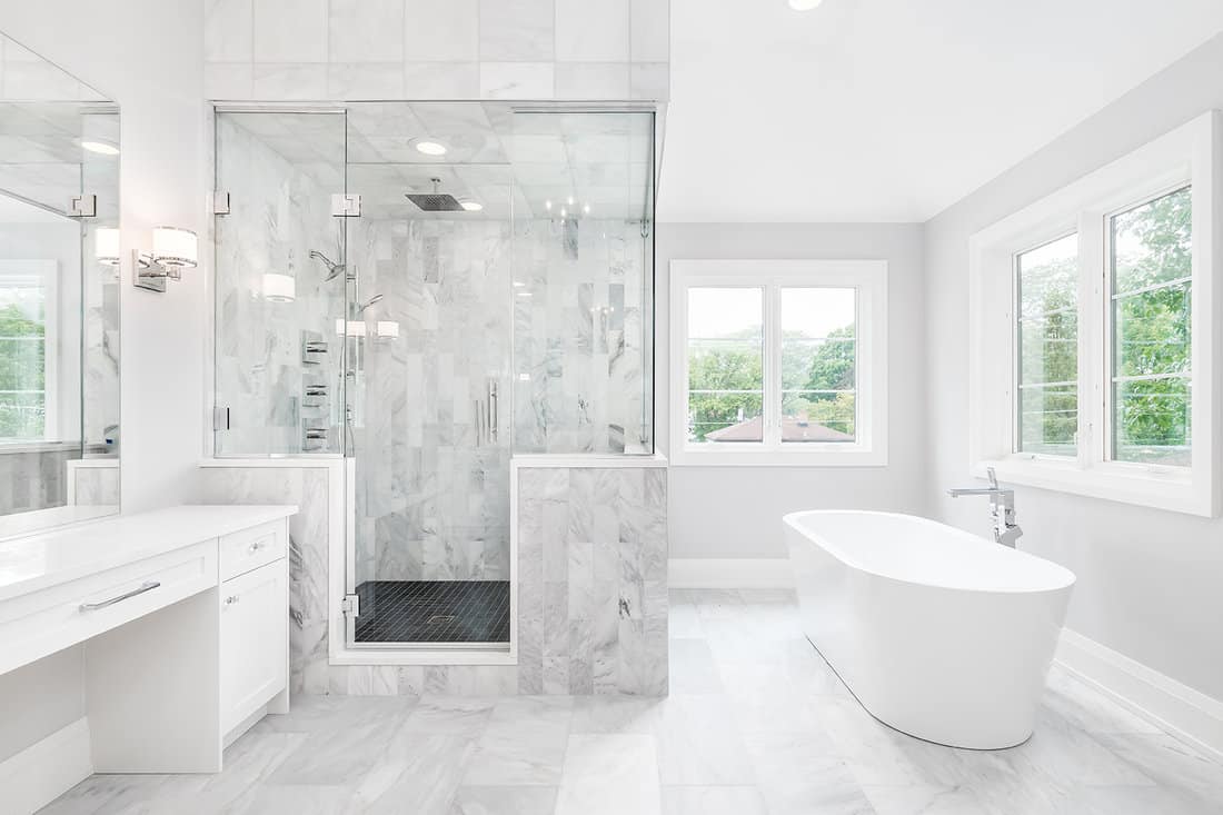 A large luxurious bathroom with a stand alone tub, white vanity, and a glass stand up shower with marble tiles and bench seat