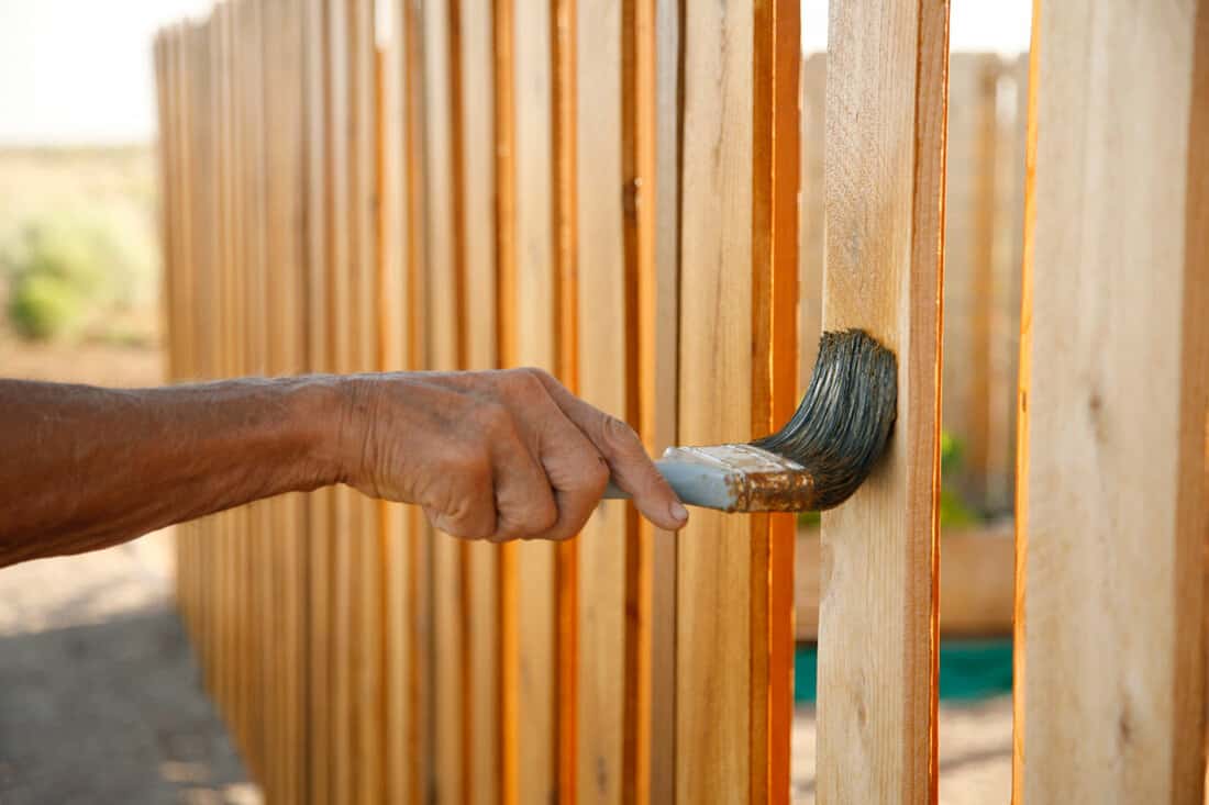 A man holding a paint brush while painting the cedar fence with wood stain